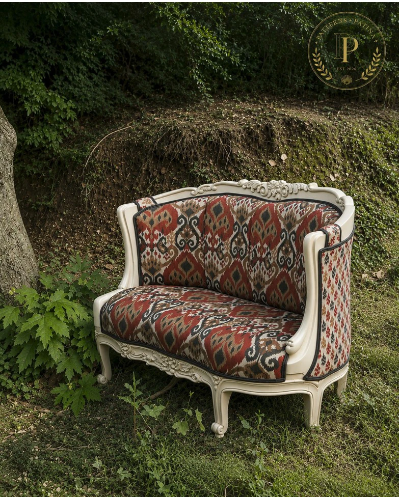 Carved cream settee in a red and black ikat, on a green bank under trees.